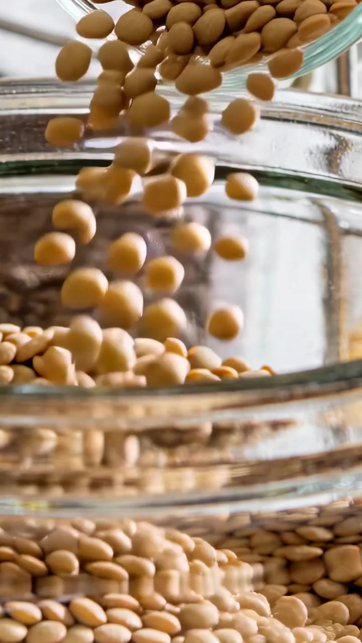 Video verticale: Contenitore che si inclina versando lenticchie marroni chiare in una ciotola sul piano di lavoro, formando un mucchio.