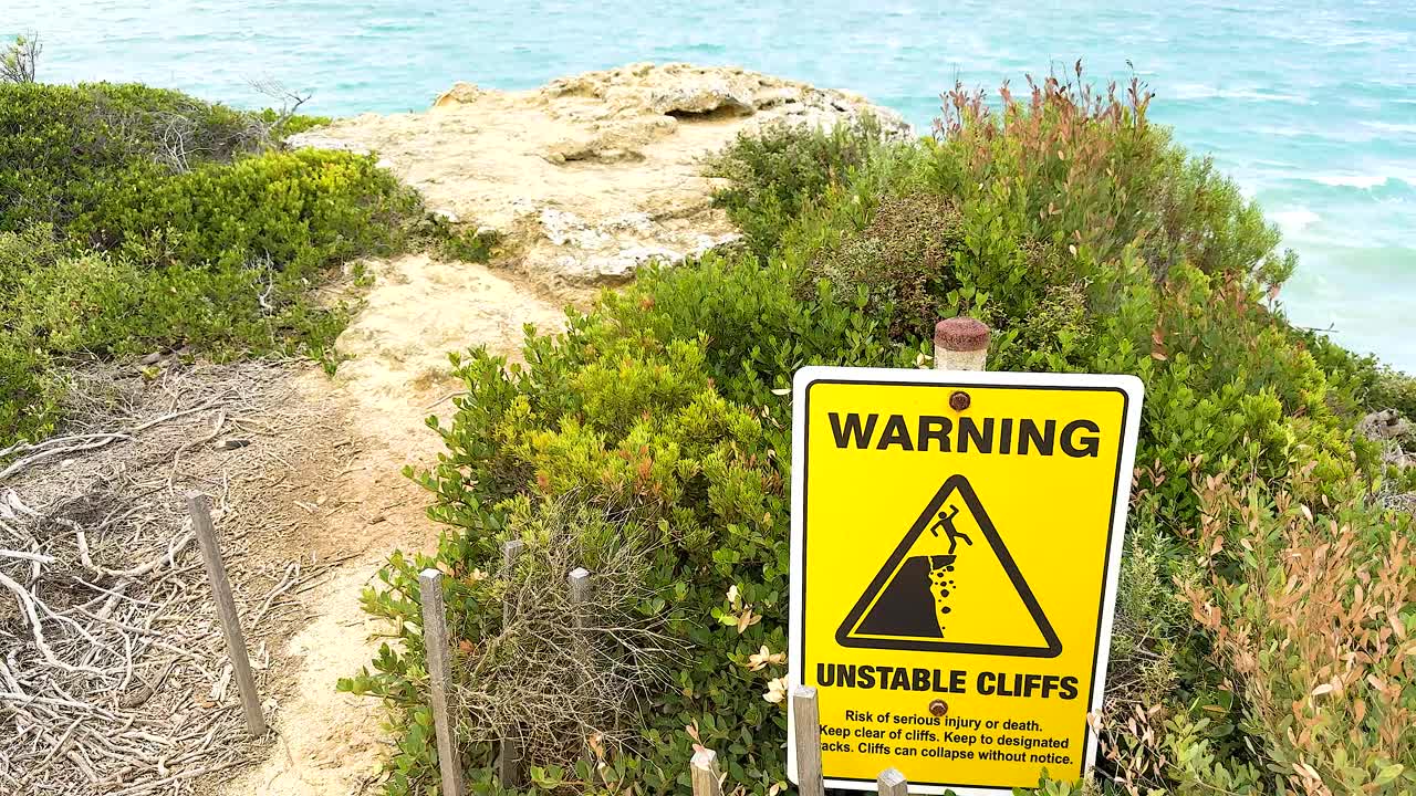 A warning sign highlights unstable cliffs along a coastal path with lush greenery and ocean views