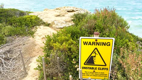 a warning sign highlights unstable cliffs along a coastal path with lush greenery and ocean views