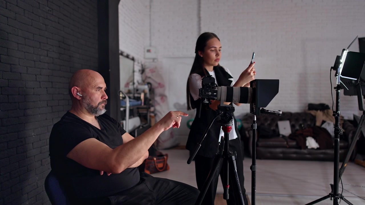 Un uomo barbuto siede su una sedia guardando il display della fotocamera. Una ragazza bruna dai capelli lunghi registra un video con il suo telefono in studio