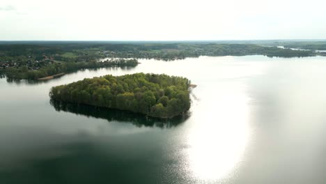 A-green-island-on-a-lake,-with-plenty-of-green-trees-on-it-and-the-mainland-visible-in-the-background