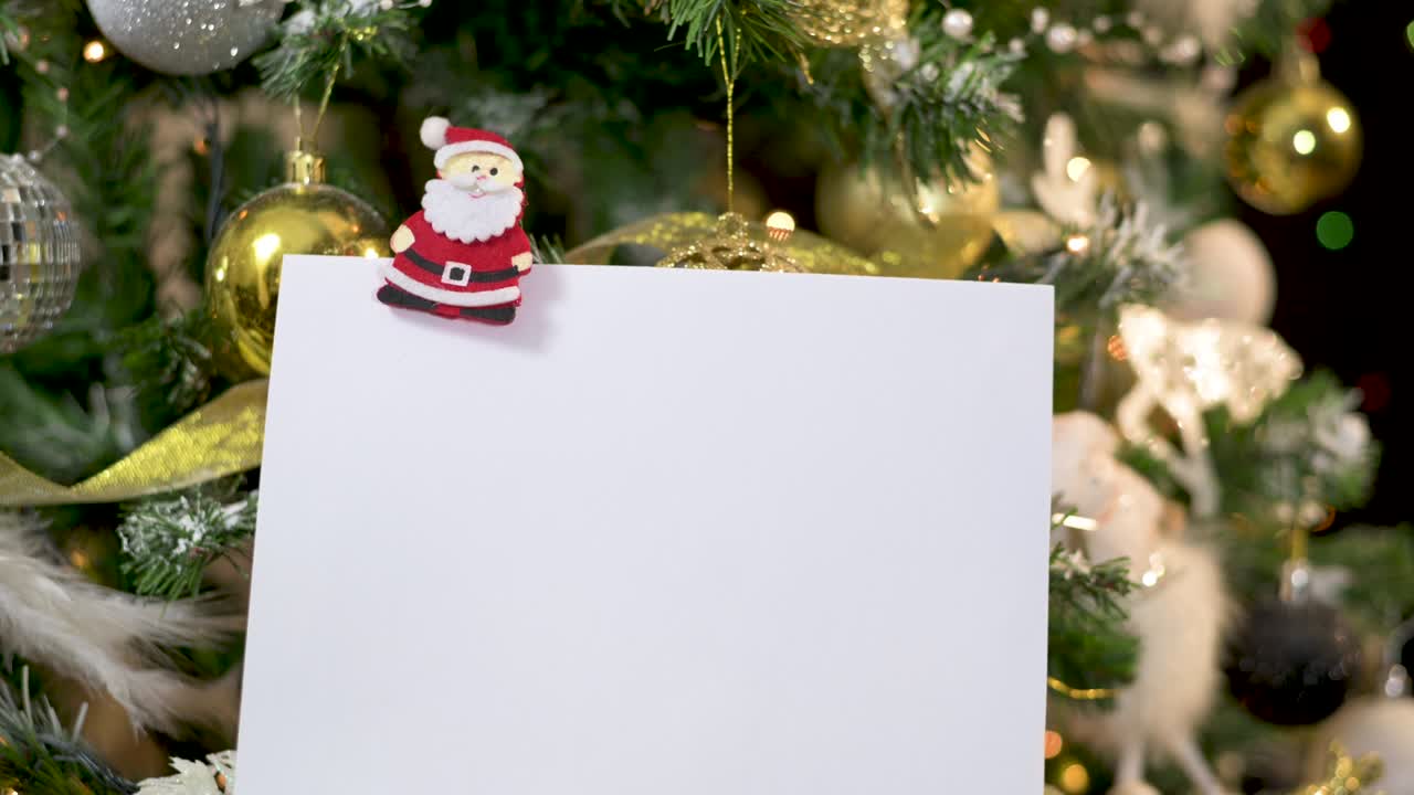cerca de la nota de tarjeta de papel enganchado con ornamento de santa en oro árbol de navidad decorado blanco con luces parpadeantes