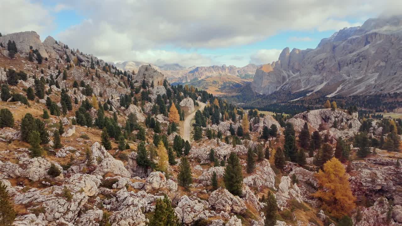 Beautiful autumn landscape in the Dolomites with a scenic view of rocky terrain and trees turning golden and orange. The winding path stretches through the colorful alpine forest below mountain peaks.