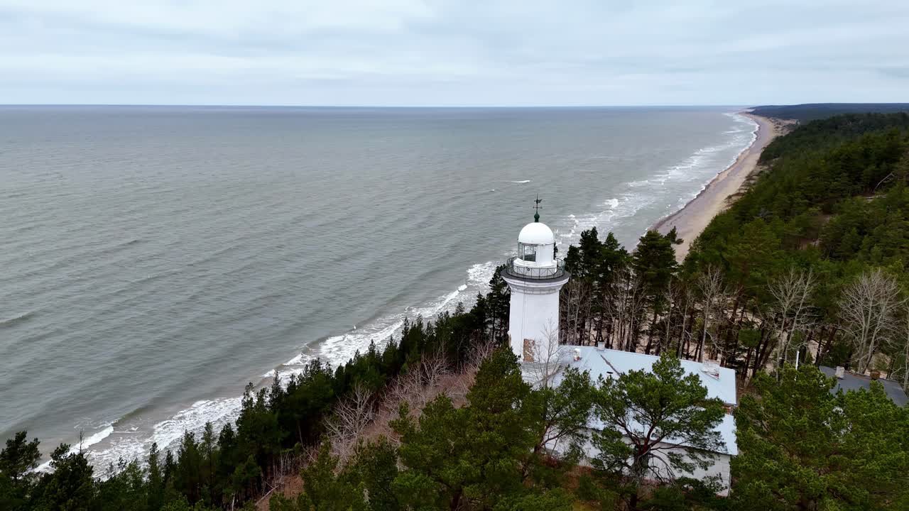 a white lighthouse with a white roof , in a green pine forest, on a grassy hill overlooking a blue ocean, Aerial Orbit Pan Shot