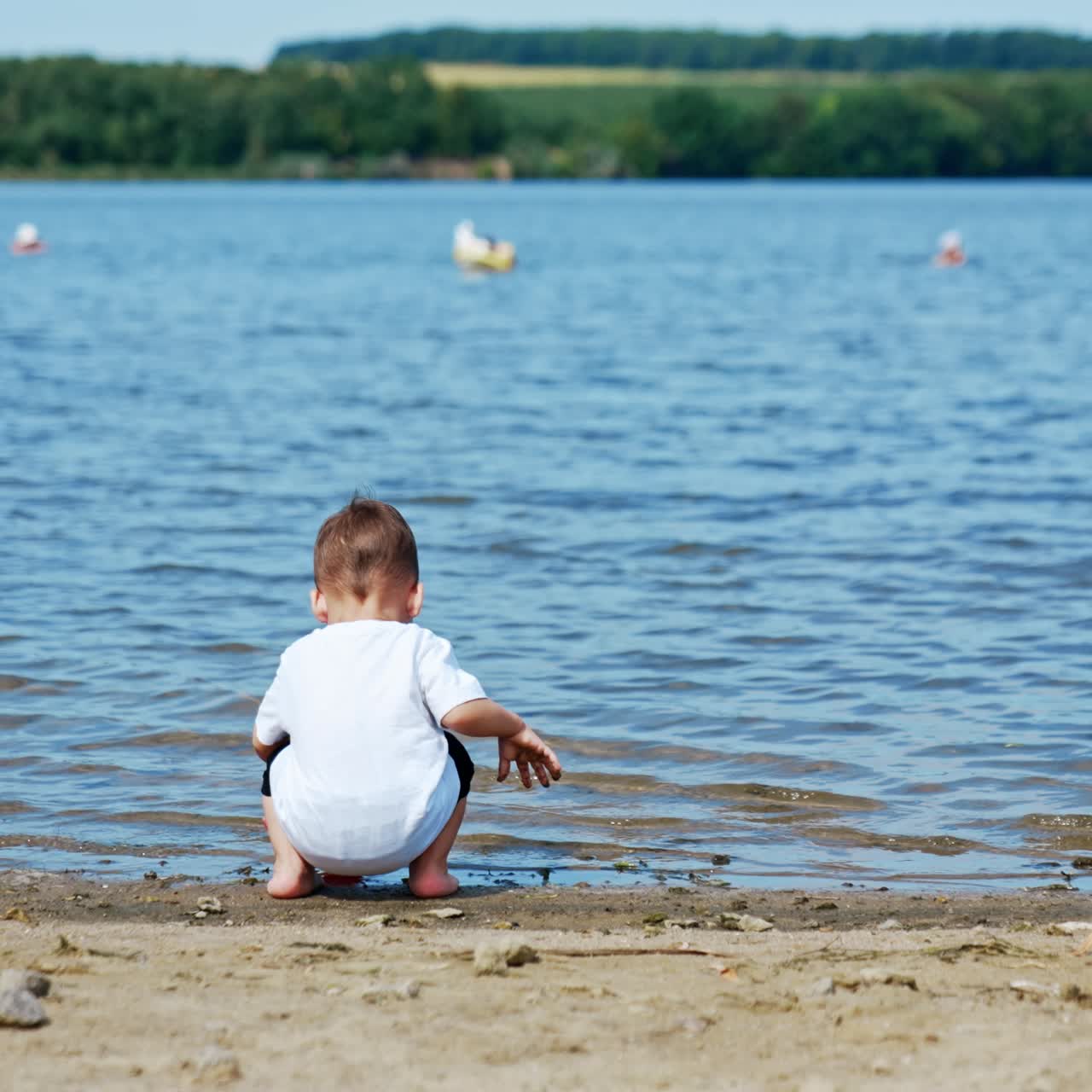 Lovely kid in white T-shirt comes up to a river. Toddler is filling his watering can with water