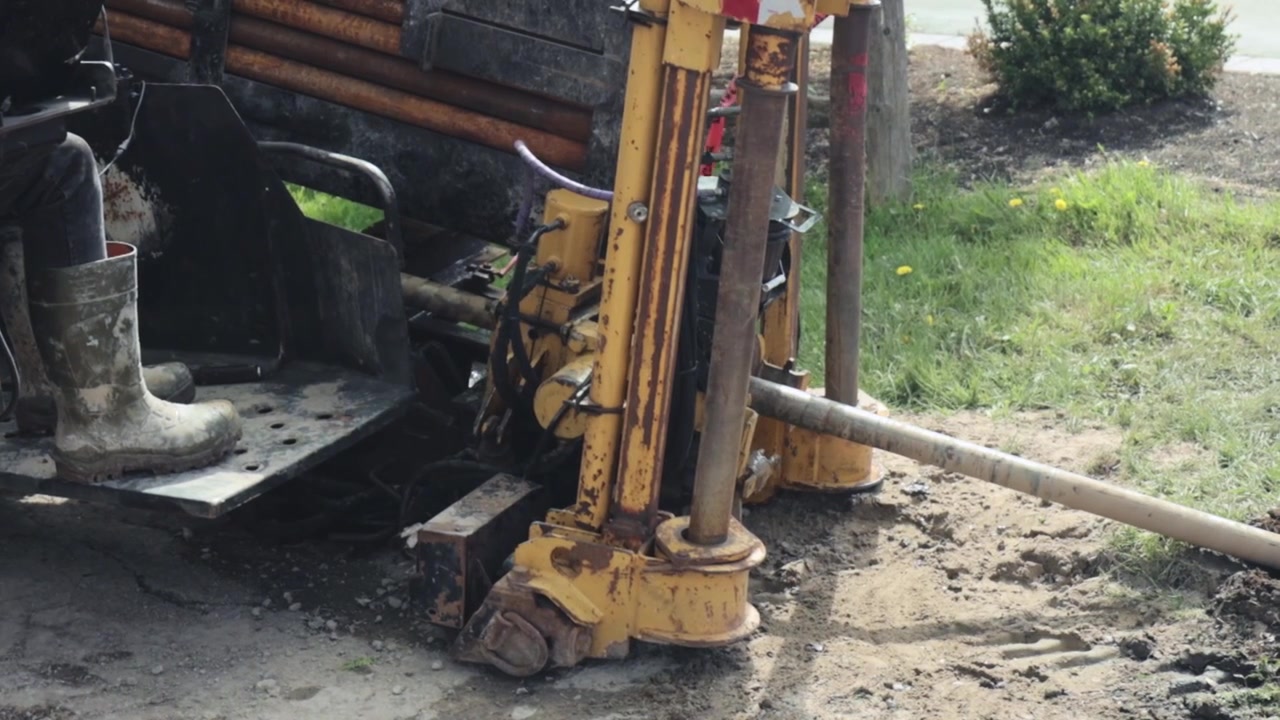 A directional drilling machine pushes a drill bit into the ground on a grassy work area, with an operator at the controls installing electrical conduit for street lighting.