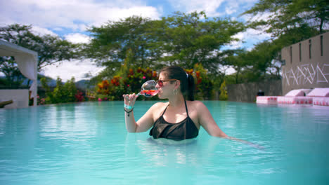 Young-woman-in-sunglasses-drinks-cocktail-in-pool,-push-in-portrait