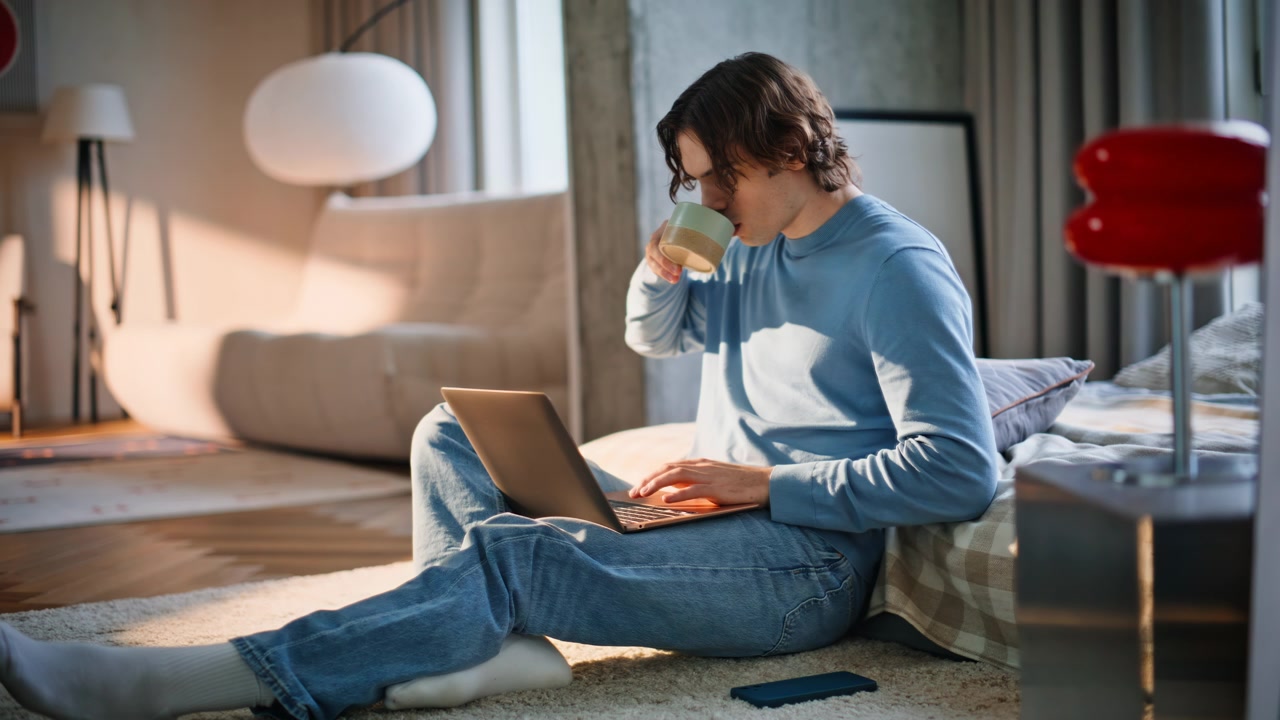 Giovane freelance che digita al laptop lavorando da remoto. Uomo rilassato che sorseggia il caffè