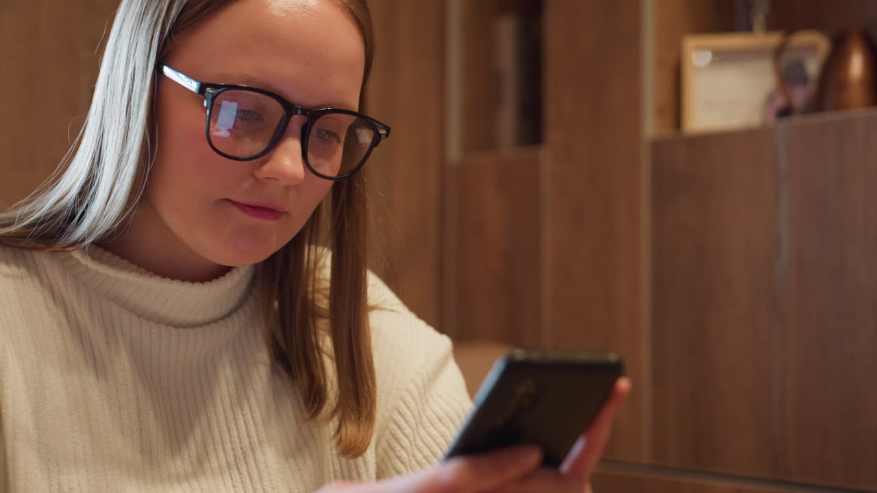 Young lady in white ribbed sweater and black glasses chatting on smartphone with visible phone screen reflection in lenses, seated in cozy wooden room with soft daylight and warm atmosphere
