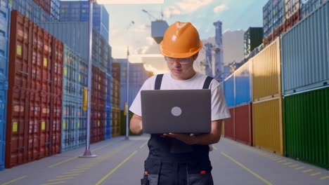 trabajador asiático con gafas de protección y casco de seguridad usando una computadora portátil mientras está de pie en el almacén del patio de contenedores