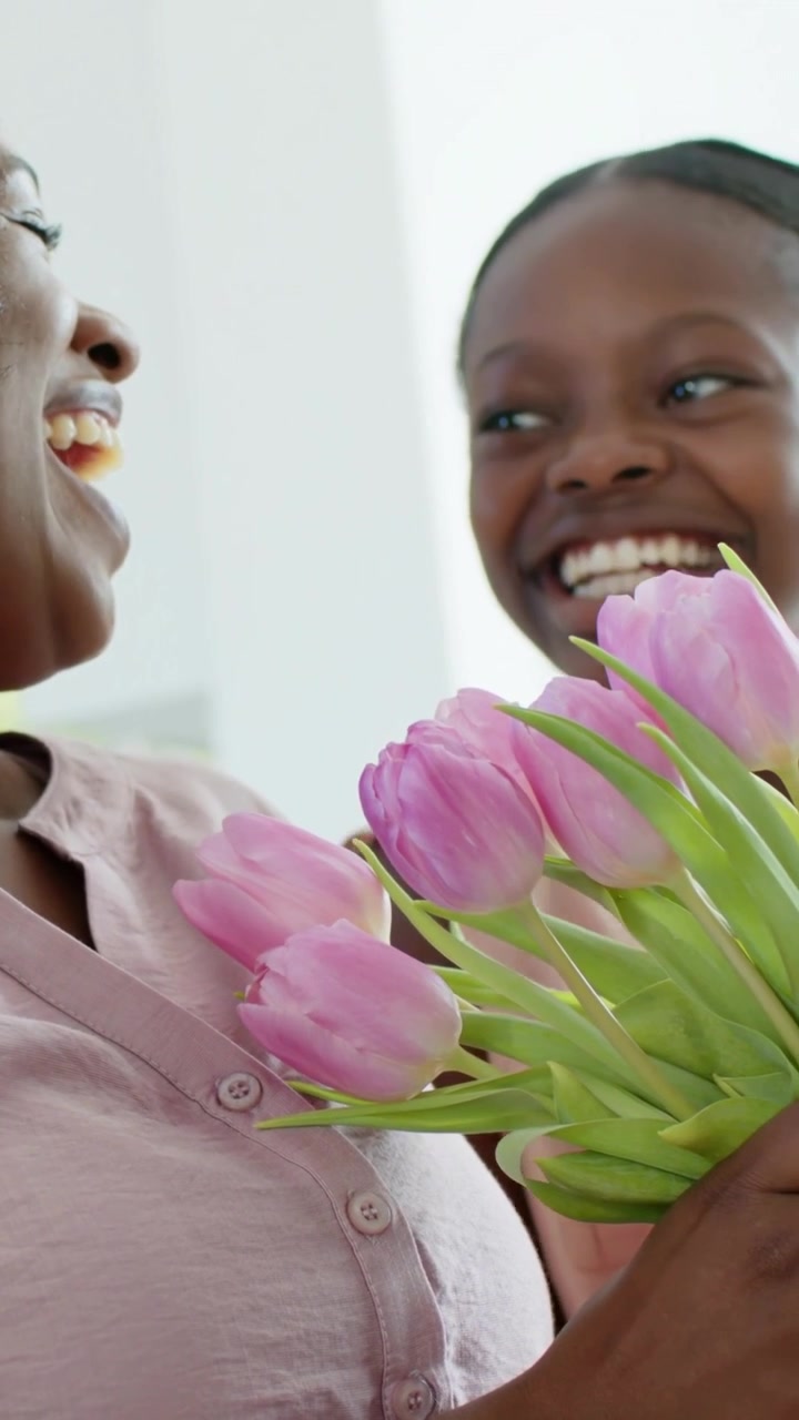 Vertical video: Entering daughter offering pink tulips to mother at home in pink tops to show love