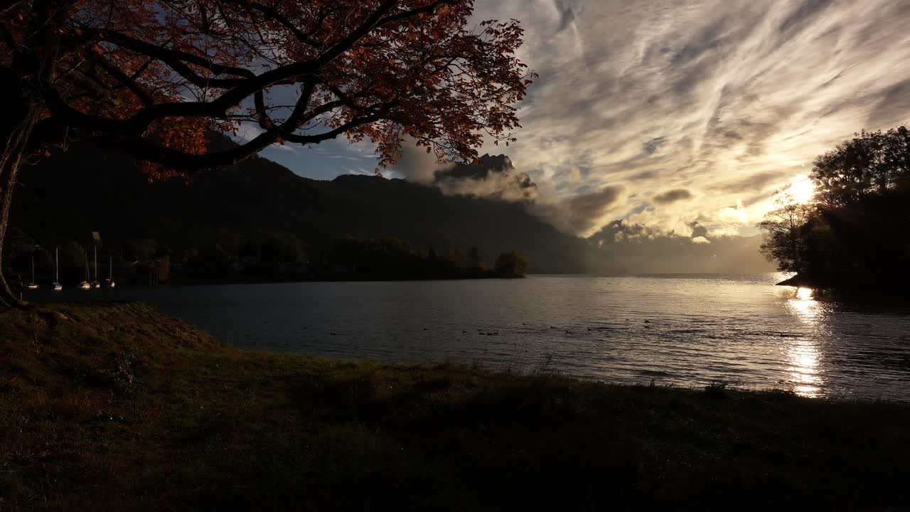 A câmera avança lentamente em direção a árvores vibrantes de outono ao lado do lago, com montanhas ao fundo e nuvens tocando seus picos durante um nascer do sol nublado com raios de sol dourados em Wessen, Amden, Glarus, Suíça.