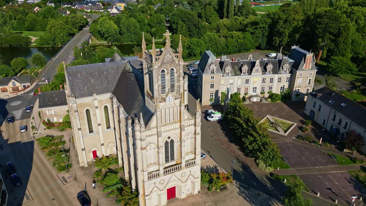 Orbiting aerial view of Changé church and town hall in Mayenne, with the river, road, and lush vegetation captured in a downward perspective - France