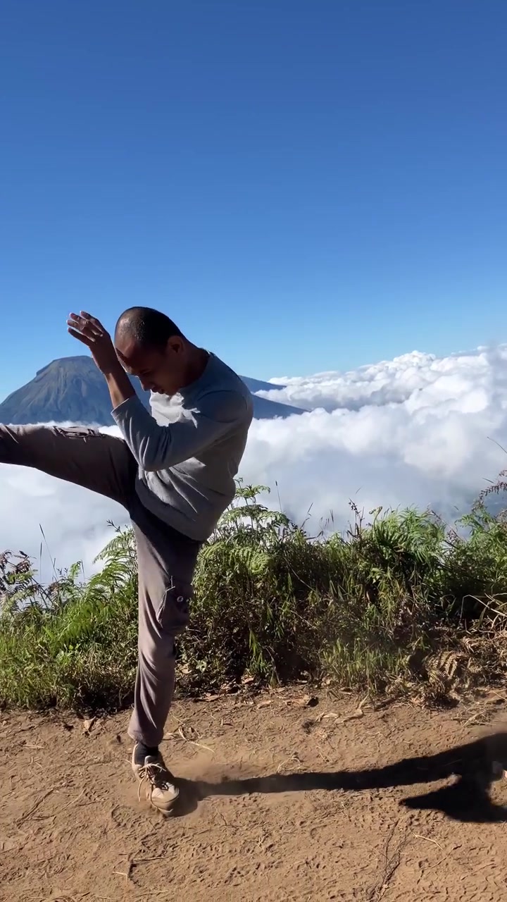 Vertical 9:16 slow motion shot of a man practicing Muay Thai shadow boxing on a mountain trail, overlooking a stunning sea of clouds and volcanic peak under a clear blue sky.