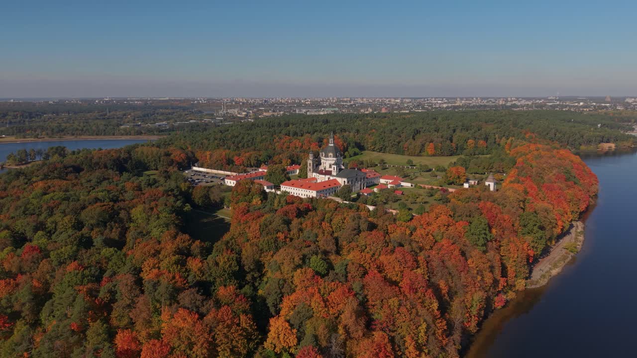 Aerial view of Pažaislis Monastery in Kaunas, Lithuania, nestled in a vibrant autumn forest by the river, with city skyline in the background under a clear blue sky, zooming in