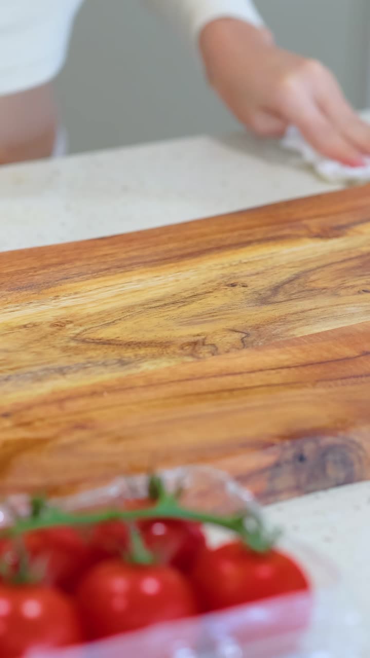 Hände wischen ein Holzschneidebrett auf einer Küchenarbeitsplatte ab, mit Tomaten in der Nähe, in einer gut beleuchteten Umgebung.