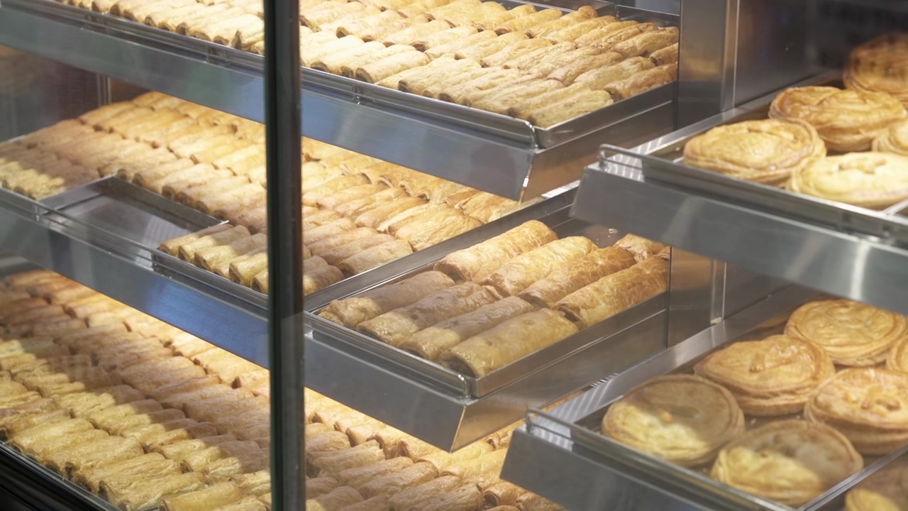 Lange brett med butikkbakte paier og pølsebrød ligger stablet i et varmeskap i rustfritt stål bak klart glass.