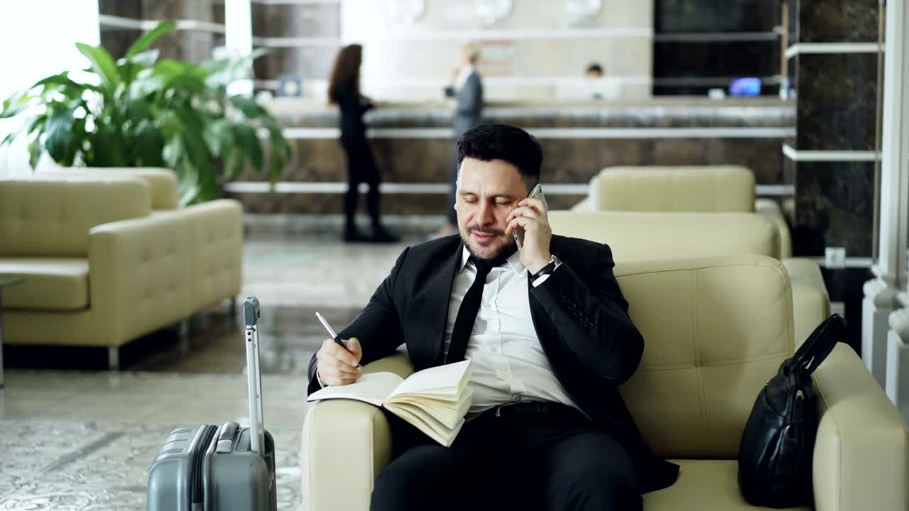 tomada de un hombre de negocios confiado y alegre hablando por teléfono móvil sonriendo y escribiendo notas en el bloc de notas mientras está sentado en el sillón en la recepción del hotel. concepto de viaje, negocios y personas