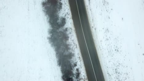 Vista-De-Pájaro-De-La-Carretera-Vacía-En-Invierno,-Paisaje-Cubierto-De-Nieve,-Coche-Negro-Conduciendo-Por-La-Carretera,-Anticipación-De-Que-Algo-Suceda