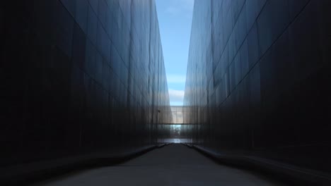 Footage between two large and tall black walls of Victims of Communism memorial during a sunny day with blue sky visible in the end and at the top. The walls are large and have reflective panels.