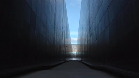 footage between two large and tall black walls of victims of communism memorial during a sunny day with blue sky visible in the end and at the top. the walls are large and have reflective panels.