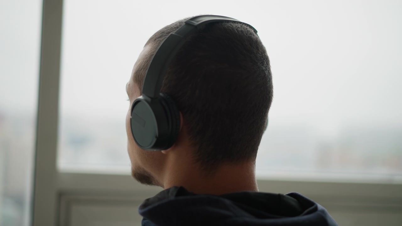 Nærbilde av mann med svarte hodetelefoner som vender mot et vindu med uskarp bybildebakgrunn. Mykt naturlig lys belyser motivet og omgivelsene, og skaper en rolig innendørs atmosfære. Mannen forblir.