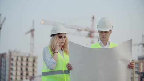 woman talking on the phone and asks the builder what is on the drawings standing on the background of buildings under construction.