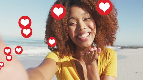 animation of red heart love digital icons over smiling woman blowing kisses on beach