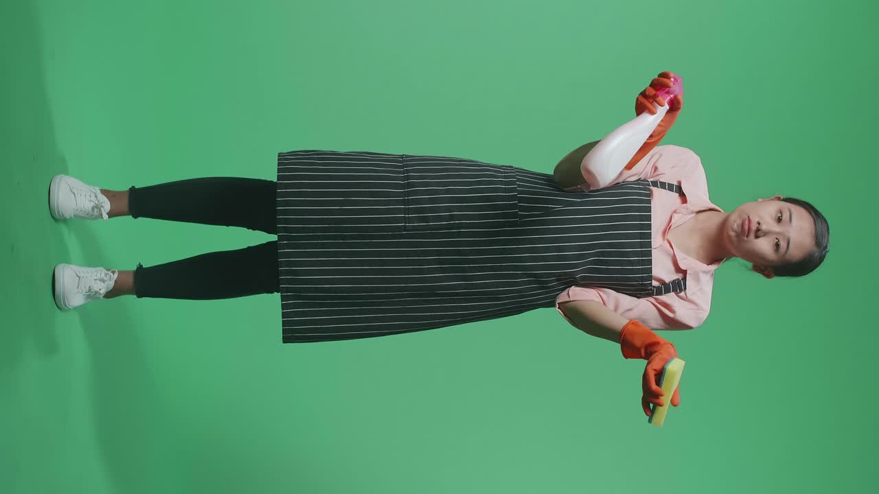 초록색 스크린 스튜디오에 서 있는 스프레이와 부 스폰지를 보여주는 전신의 혼란스러운 아시아 여성 집주인