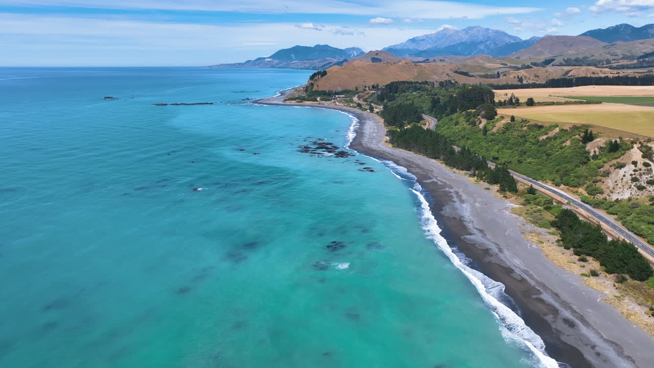Aerial flies south above the aqua Pacific Kaikoura Coast, following Black Beach towards the settlement of Kekerengu, on a hot fine day with vast mountains behind New Zealand Aotearoa
