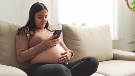 Eine-Junge-Schwangere-Hispanische-Frau-Sitzt-Mit-Dem-Smartphone-Auf-Der-Couch