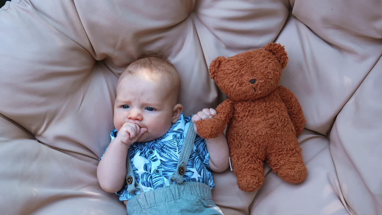 Peaceful cute infant boy in blue shirt lies chewing his fist. Adorable baby boy holding a teddy. Close up.