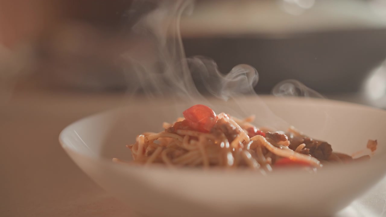 Un primo piano da angolazione bassa mostra spaghetti fumanti impiattati in una ciotola bianca poco profonda con guarnizione di pomodoro, salsa lucida, illuminazione laterale calda, ombre morbide e una presentazione invitante di un pasto appena servito.