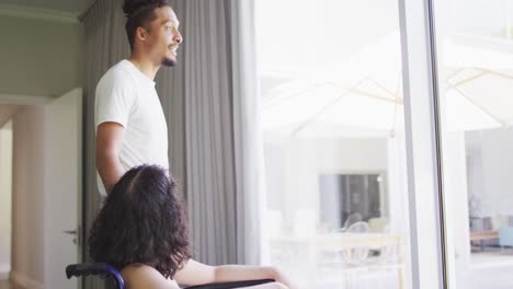 Biracial-woman-in-wheelchair-and-smiling-male-partner-looking-out-of-living-room-window