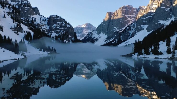 L'aria fresca del mattino provoca una nebbia bassa che si diffonde su un lago alpino centrale, riflettendo le cime illuminate dal sole.