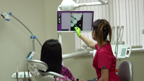 Young-female-doctor-pointing-on-the-screen-with-x-ray,-explaining-her-patient-needed-treatment.-Young-woman-sitting-in-the-dentist-chair-and-looking-at-the-monitor.-Shot-in-4k