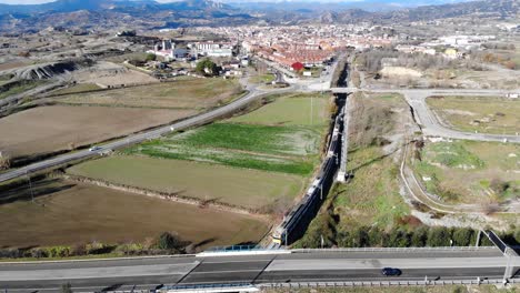 Aerial:-regional-train-passing-under-a-main-road-and-entering-a-medium-sized-town-in-Catalonia-with-the-Pyrenees-mountain-range-in-the-background