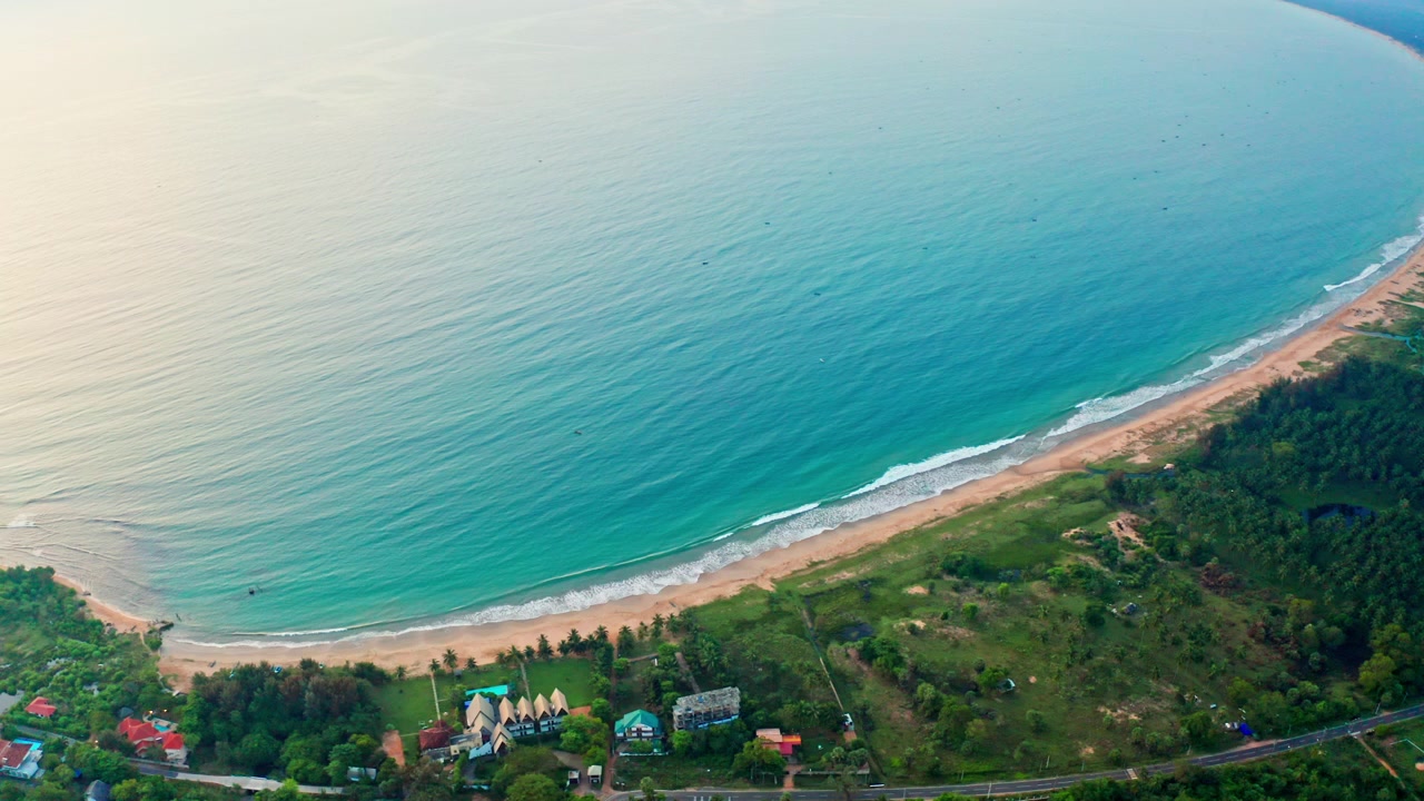 Vista aérea de una extensa playa en forma de media luna que se une al océano turquesa, un pueblo costero escondido entre palmeras bajo un suave cielo vespertino en tonos pastel