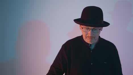 close-up view of a man in a black shirt, black hat, and glasses standing against a white background with a soft gradient of blue and pink light, creating a thoughtful and introspective mood