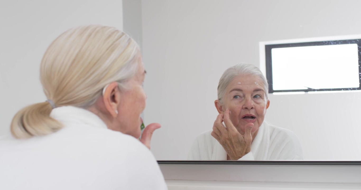 Donna anziana in accappatoio bianco alla toeletta, si guarda allo specchio mentre apre un vasetto e applica la crema idratante.