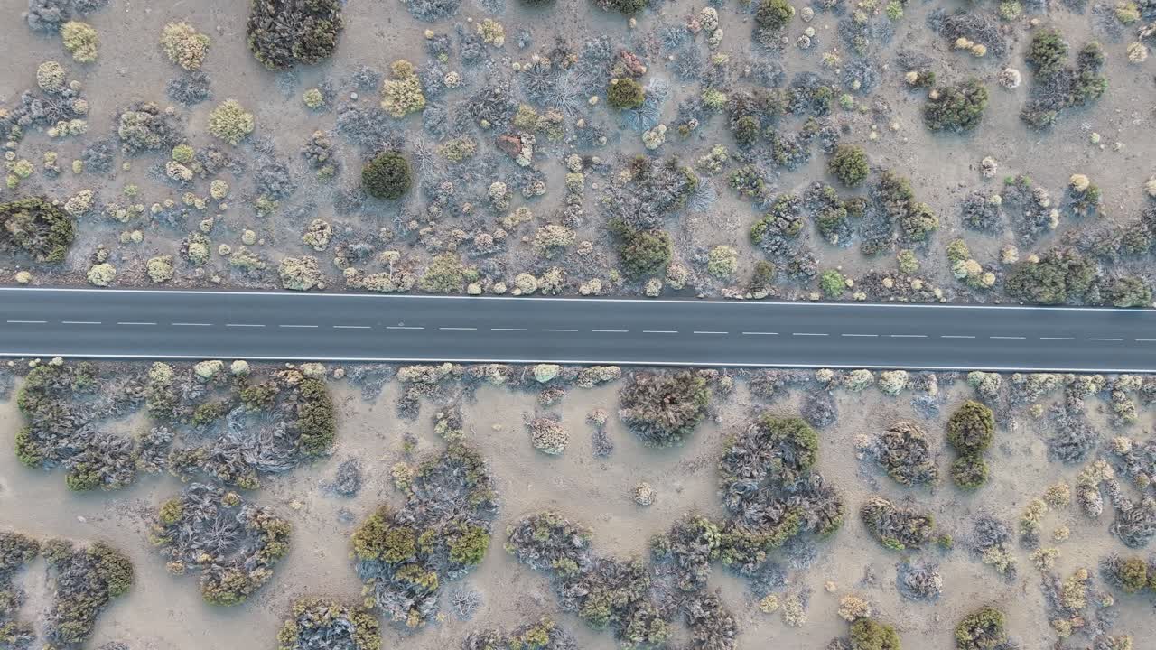 Drone flying over straight road through arid volcanic terrain with scattered shrubs