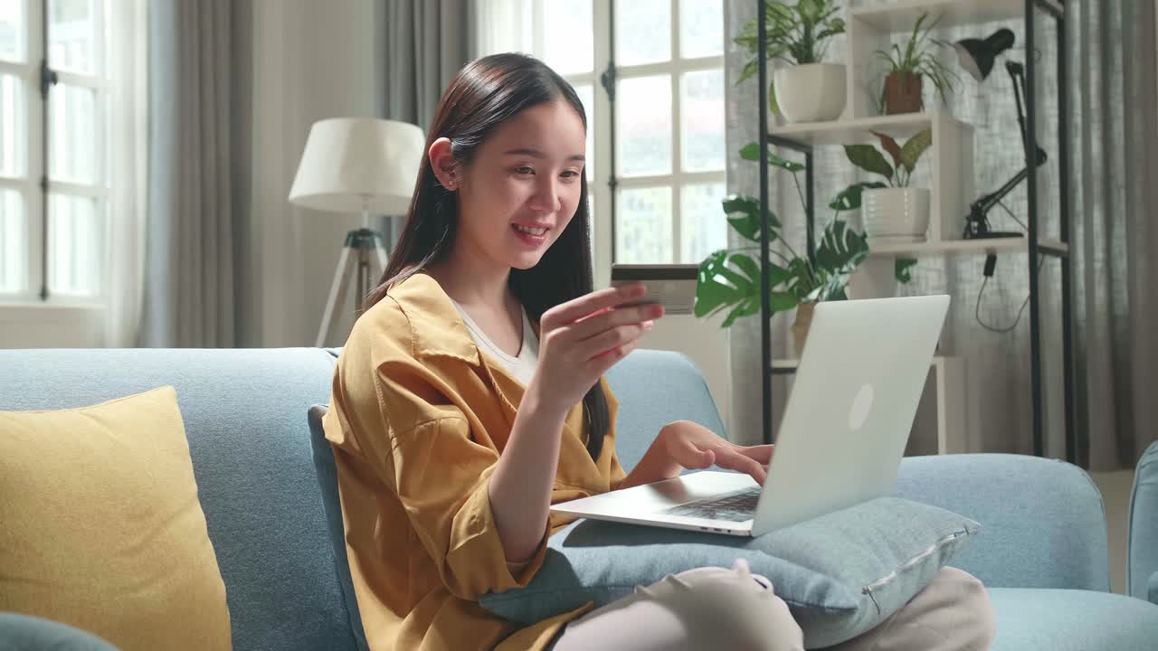 Happy Asian Woman Sitting On Couch In Living Room Holding Credit Card And Using Laptop Computer Shopping Online