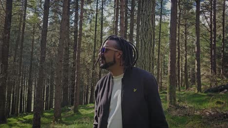 young black man wearing sunglasses walking in forest, looking at scenic nature, while hiking alone on summer day in ireland