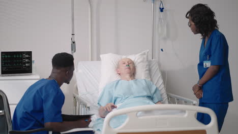 african young doctor team consulting covid-19 infected patient in bed at hospital. sick man lying in hospital bed and talking to surgeon in scrubs and protective mask. doctor examining elderly patient