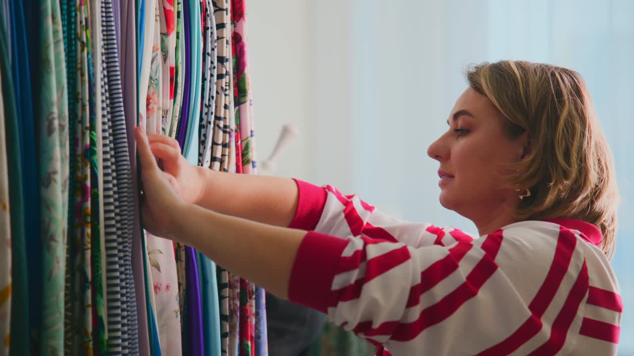 Woman selects fabric from a colorful rack. She touches materials with focused attention. Her expression shows calm interest in textiles. Suitable for fashion, crafting, or retail project scenes