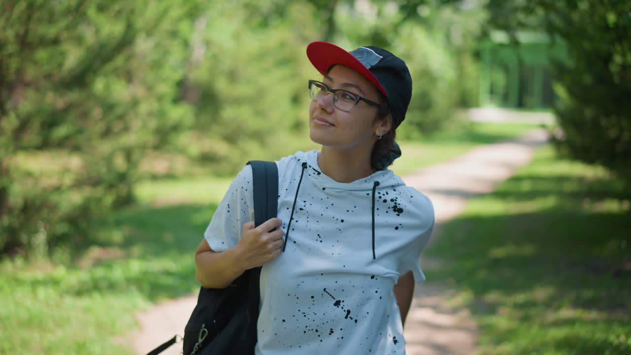 Retrato de una persona ajustándose la gorra en un sendero con una sonrisa pícara y gafas que reflejan la luz, con un aire de creador e influencer, pose segura con sudadera informal y mochila, parque verde de fondo, calidez veraniega