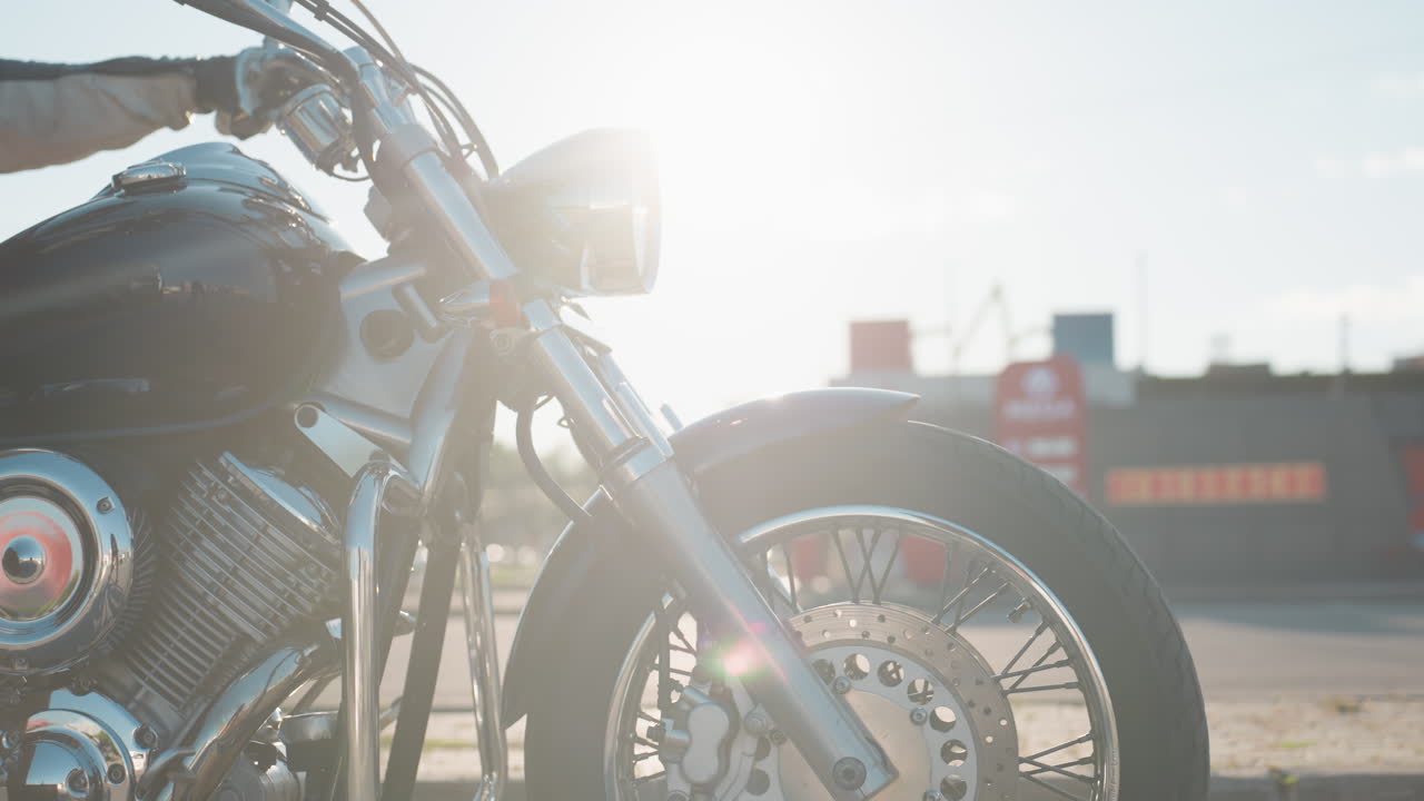 Close up of woman stepping onto motorcycle with rider holding handlebars, chrome headlight gleaming under sunlight, both wearing casual biker gear on open city street during daytime ride