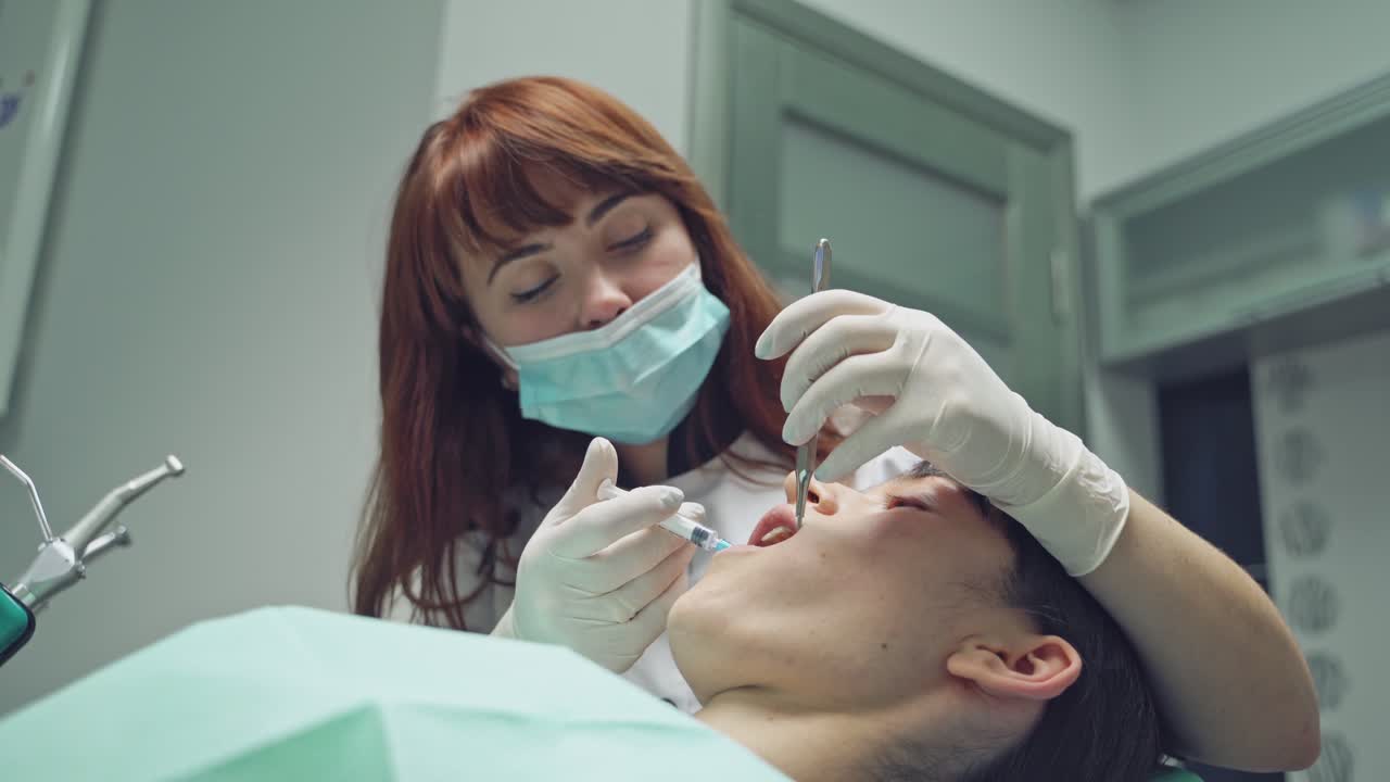 Female stomatologist applying anesthetic injection before oral surgery operation in clinic. Pretty dentist using mouth mirror and medicine syringe during her work. Close-up