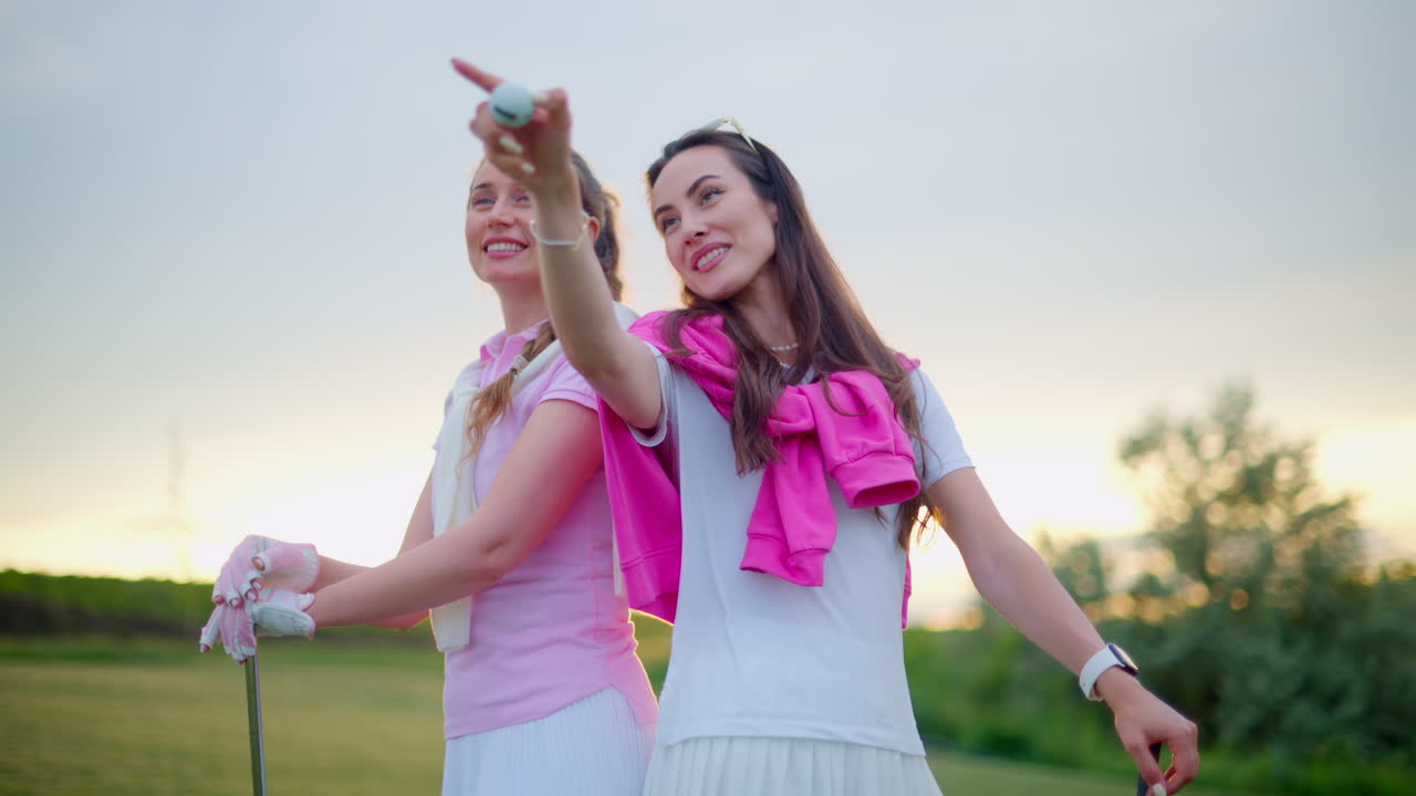 Two women in white and pink clothes, standing back to back with golf clubs in their hands, on a grass field