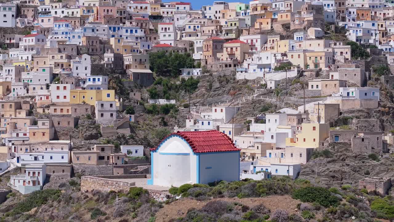 Drone orbit around a whitewashed chapel in Karpathos reveals the colorful village of Olympos in the background. Stunning aerial view of Greek island charm and mountain scenery